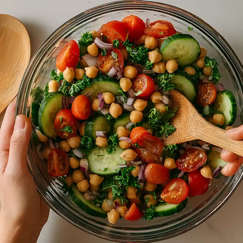 Une main mélange une salade colorée dans un grand saladier en verre, remplie de concombres, tomates, pois chiches, oignons rouges et persil haché.