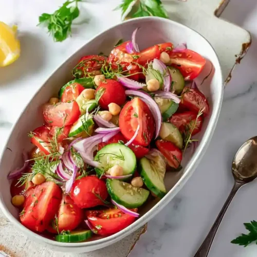 Une salade de concombre et tomate fraîchement servie dans un grand plat, garnie d’oignons rouges, pois chiches et herbes fraîches, posée sur une table ensoleillée.