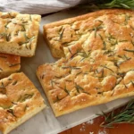 Focaccia maison moelleuse garnie de romarin, découpée en carrés dorés et croustillants, parfaite pour un déjeuner italien convivial.
