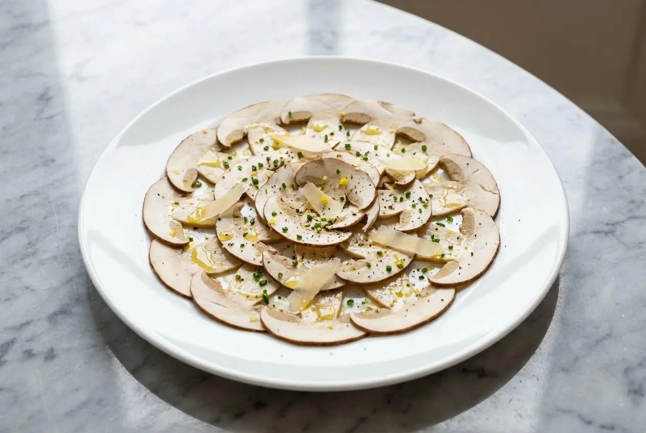 Gros plan sur un carpaccio de cèpes, les fines tranches de champignon cru arrangées en rosace, rehaussées de zestes de citron et d'un filet d'huile de noisette brillante sur une assiette en céramique minimaliste.