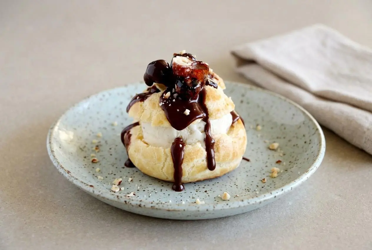 Profiteroles au chocolat empilés dans un bol blanc, nappés d'une sauce chocolat brillante et garnis de noisettes caramélisées.