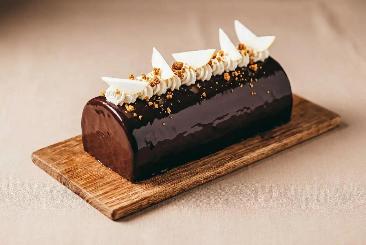 Bûche poire Spéculoos entière avec un glaçage miroir au cacao brillant, décorée de brisures de spéculoos et de petites meringues.