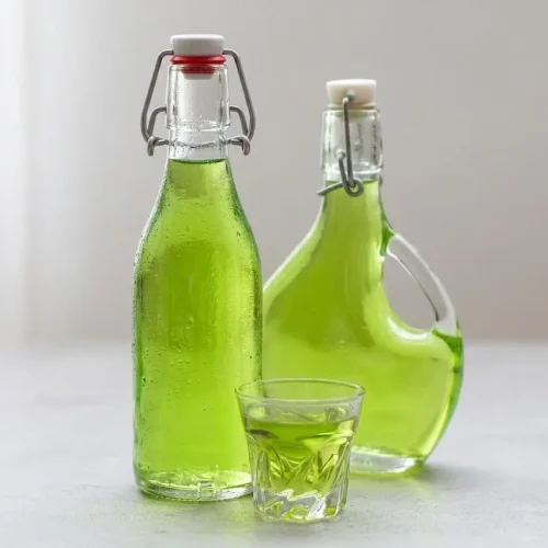 Une bouteille de Liqueur de Verveine maison avec deux verres remplis de la liqueur verte brillante, accompagnés de feuilles de verveine