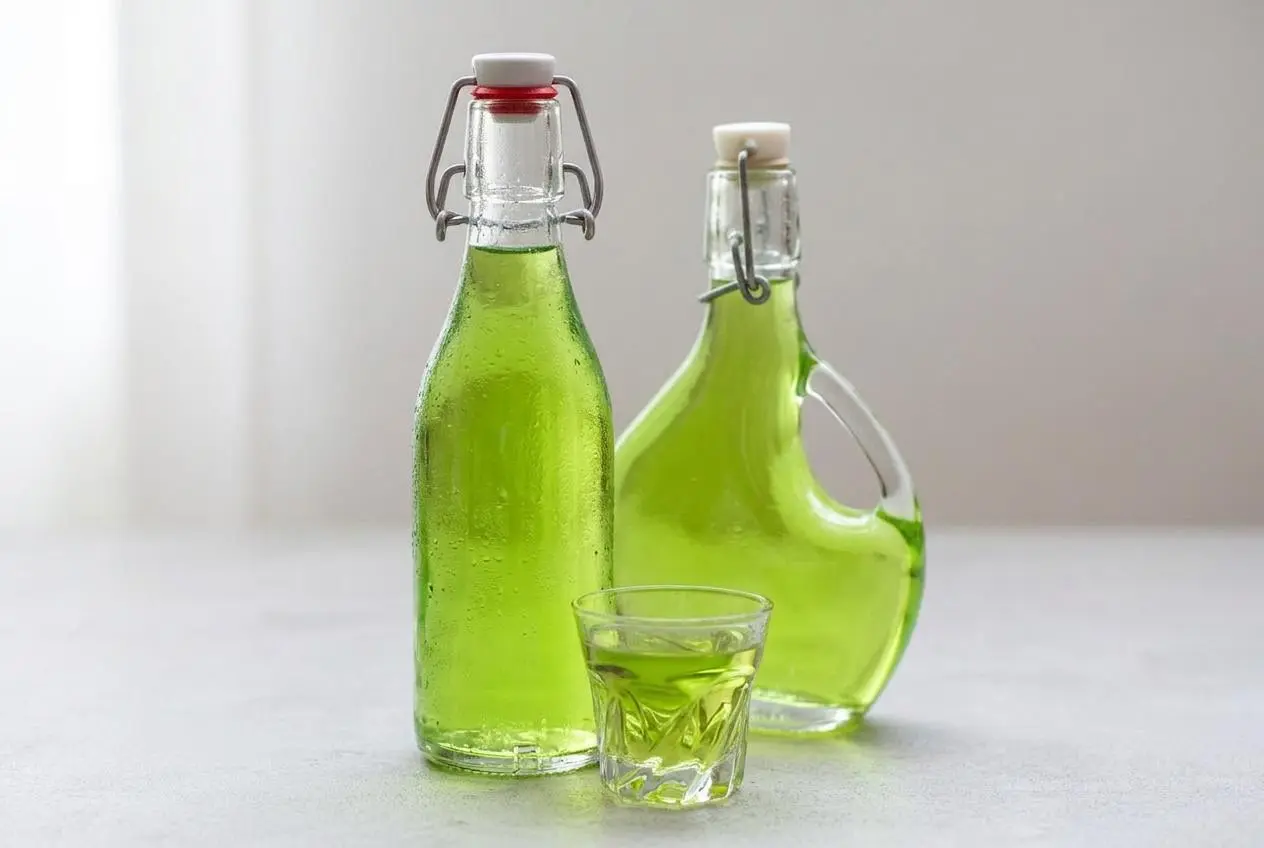 Une bouteille de Liqueur de Verveine maison avec deux verres remplis de la liqueur verte brillante, accompagnés de feuilles de verveine