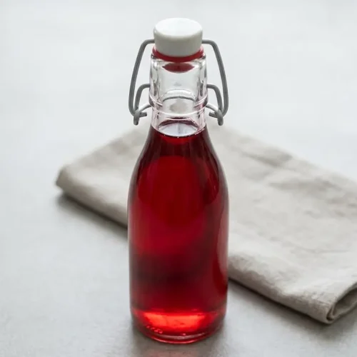Bouteille en verre de sirop de fraise maison, d'une couleur rubis intense, à côté d'un verre et de fraises fraîches.