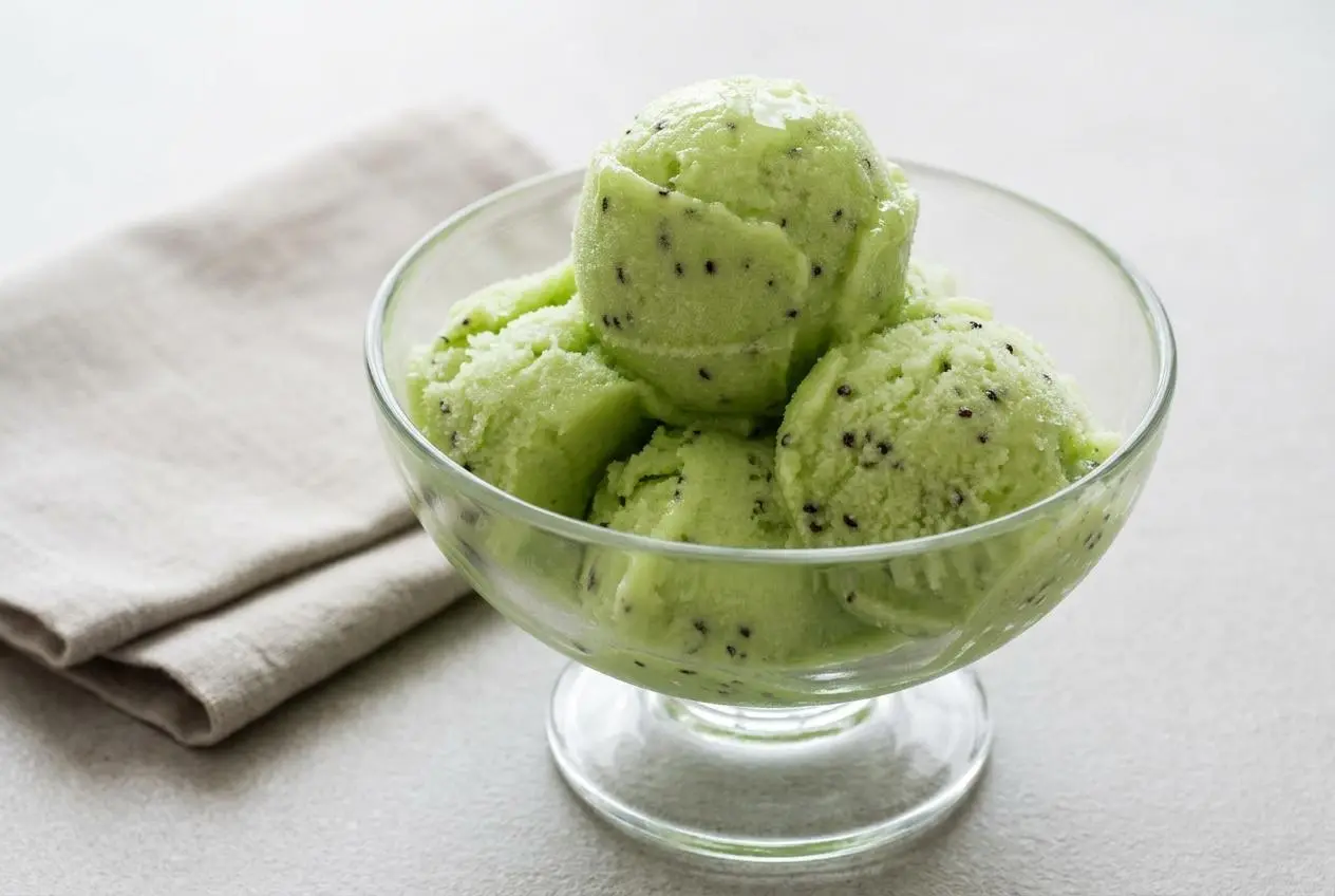 Deux boules de sorbet au kiwi vert pâle dans un bol blanc, garnies d'une feuille de menthe, avec des kiwis frais à côté.